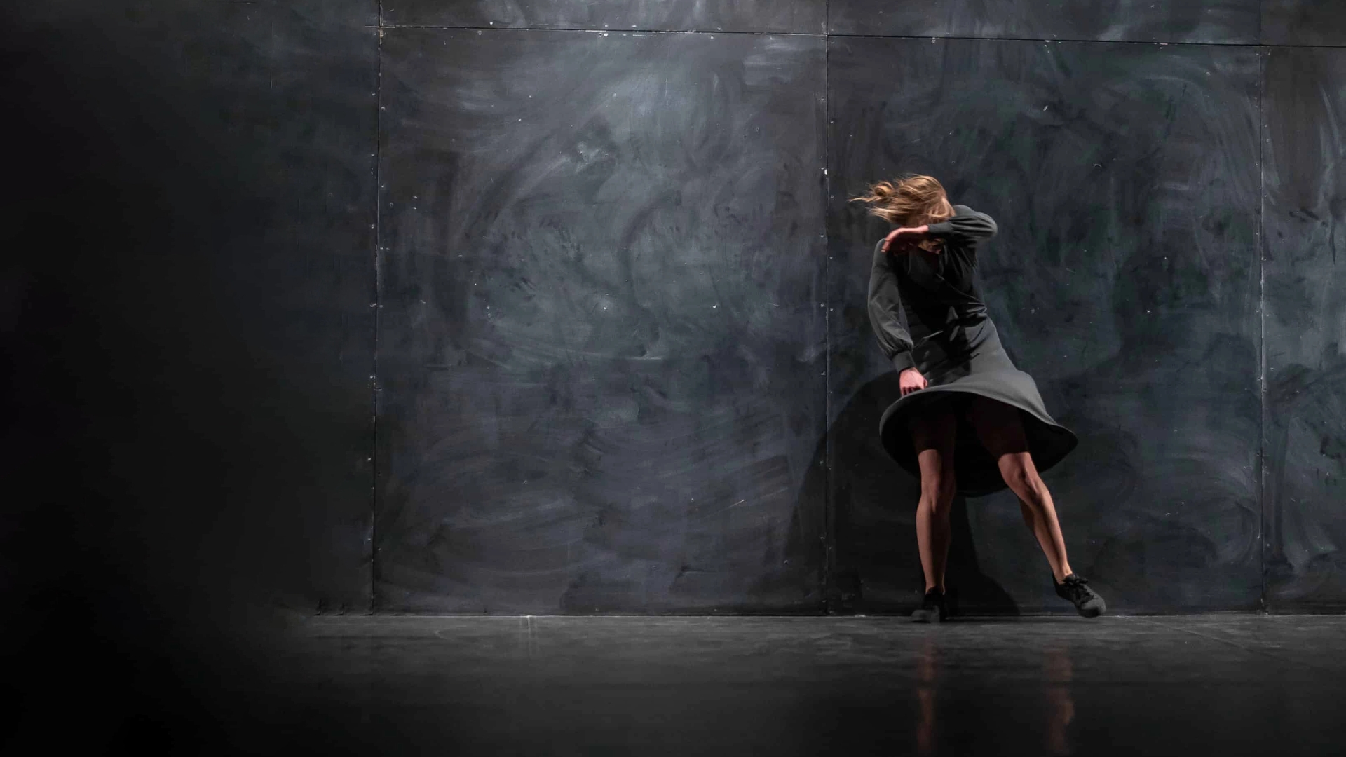 A person in a dark dress is caught mid-motion against a dark background, with their hair flying and arm raised, suggesting energetic movement.