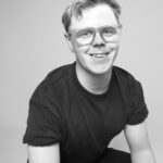 A smiling person with short hair and glasses wears a dark T-shirt in a black and white portrait. The image conveys a friendly and approachable tone.