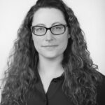 A woman with long, curly hair is wearing a dark button-up shirt, set against a plain white background in a monochrome style.