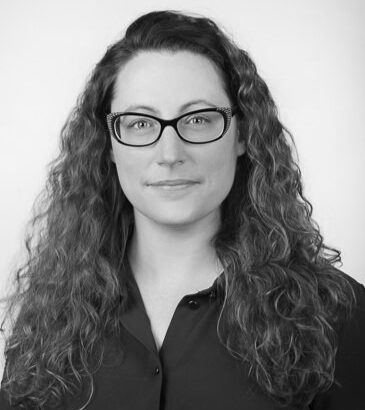 A woman with long, curly hair is wearing a dark button-up shirt, set against a plain white background in a monochrome style.