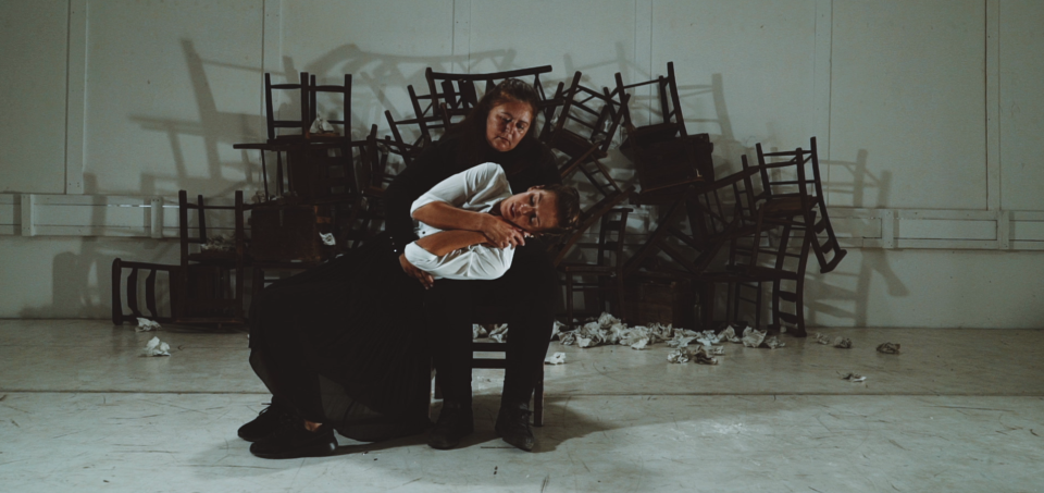 Two performers sit on a chair in an embrace, one cradling the other, who leans in with eyes closed. Behind them is a chaotic pile of overturned chairs and scattered paper, set against a stark white wall.