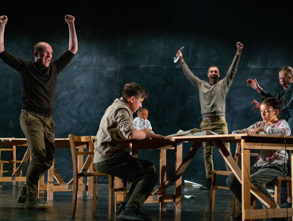 A lively stage scene with five actors celebrating, two seated at a table and three standing, conveying excitement and camaraderie.
