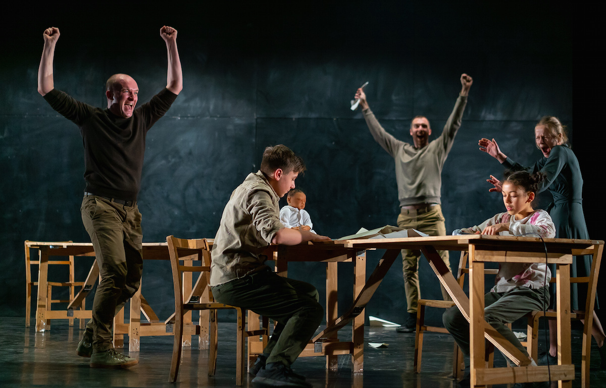 A lively stage scene with five actors celebrating, two seated at a table and three standing, conveying excitement and camaraderie.