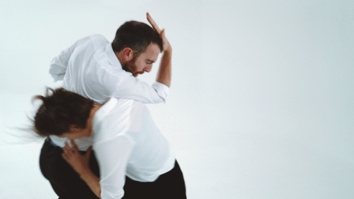 A man and woman in white shirts perform a contemporary dance against a plain backdrop, their movements dynamic and fluid, expressing grace and intensity.