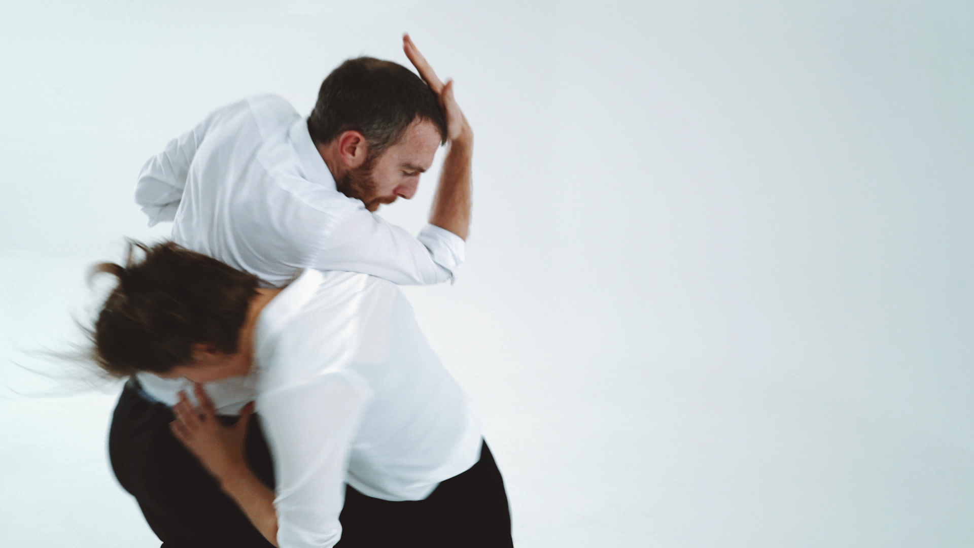 A man and woman in white shirts perform a contemporary dance against a plain backdrop, their movements dynamic and fluid, expressing grace and intensity.